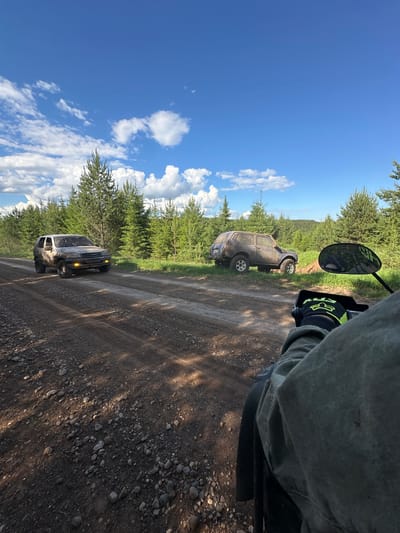 The image shows a dirt road with two off-road vehicles parked on either side of the road. A motorcycle, the point of view for the picture, is on the road. The location is presumably a rural area with trees lining the road. The sky is blue with some clouds. There are no people visible besides the possible rider of the motorcycle. The state and specific location within the city of Пепеляева, Russia, are not apparent.