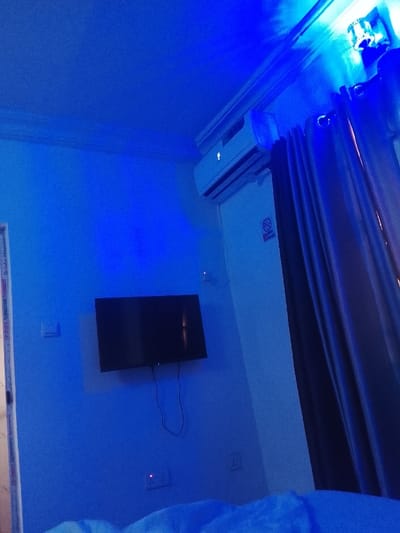 A room bathed in blue light reveals a wall-mounted flat-screen TV with dangling cords. An air conditioning unit sits near the ceiling above a "no smoking" sign. Shiny, dark curtains hang to the right. Bedding is visible in the foreground, all cast in the same blue hue. A single electrical outlet is visible below the TV.