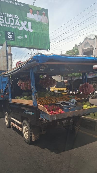 A black pickup truck, modified into a mobile fruit stand, is parked on the side of a paved road in Pantegajah, Indonesia. The truck is laden with a variety of fruits, including watermelons, oranges, and dragon fruit. A blue tarp covers the top of the stand, and red bags of merchandise hang from the structure. The truck's license plate reads "BL 8157 YB". In the background, a yellow Fuso truck and motorcycles are visible. Above the fruit stand, a large green billboard displays images of men in traditional Islamic attire and text in Indonesian. The sky is clear and bright.