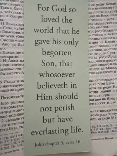 The image shows a close-up of two pages of text, one in English and one in Bulgarian, that appear to be from a religious text. The English text, in a large, bold font, reads "For God so loved the world that he gave his only begotten Son, that whosoever believeth in Him should not perish but have everlasting life. John chapter 3, verse 16." The Bulgarian text is in a smaller font and appears to be a biblical passage as well, likely related to genealogies or historical accounts based on the visible words like "роди", "синове", and names. The pages are slightly creased and show some wear. The background is a light, neutral color. There is no visible indication of the city of Newry, United Kingdom, or any specific location within it.