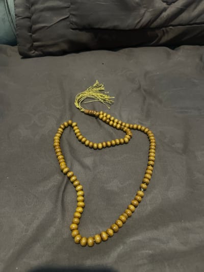 A string of wooden prayer beads, commonly known as tasbih, is laid out on a dark brown, wrinkled fabric surface. The beads are light brown and oval-shaped, strung together in a loop. A small, golden tassel is attached to one end of the string. The background consists of a dark, textured surface that appears to be a bed or sofa. The lighting is dim, casting soft shadows. The image is taken from a slightly elevated angle, looking down at the prayer beads. There are no people or animals visible in the image. The location is not discernible from the image itself and cannot be confirmed as Bandarbireuen, Indonesia.