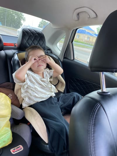 A young child is seated in a car seat in the back of a vehicle, looking tired with their eyes covered by their hands. The child is wearing a white patterned shirt and dark pants. In the background, a building with a blue roof is visible through the car window, alongside trees and a road. The interior of the car, including the seats and headrests, is visible. The scene appears to be within the city of Kushnarenkovo, Russia.