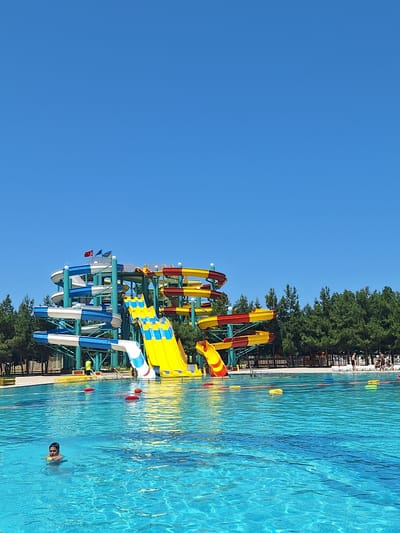 The image shows a water park on a clear, sunny day. A large, multi-colored water slide structure dominates the background, with various tubes and chutes in shades of blue, white, yellow, and red. Two flags are visible at the top of the structure. In the foreground, a bright blue swimming pool is filled with water, with several yellow and red floating buoys scattered on the surface. A young boy wearing a yellow life vest is swimming in the pool, holding a yellow object. In the background, beyond the pool, there is a line of green trees. Further in the distance, a group of people are visible near lounge chairs and a fence. The scene appears to be a recreational area, likely a popular spot for families and individuals seeking outdoor activities. The location is specified as the city of Khudat, Azerbaijan.