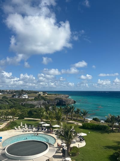 An elevated panoramic view displays a tropical coastal landscape in Wellhouse, Barbados, under a bright blue sky with scattered cumulus clouds. The scene encompasses a resort area in the foreground transitioning to natural cliffs and the Atlantic Ocean.
In the lower foreground, a curvilinear swimming pool with light blue water contains approximately four individuals. Adjacent to it, a circular feature with a dark cover is present. Surrounding the pool on a light-colored paved deck are numerous white lounge chairs, some with blue cushions, and several white market umbrellas. Approximately seven individuals are visible lounging on these chairs or standing nearby. Well-maintained green lawns, interspersed with numerous palm trees and tropical foliage, extend from the paved pool area towards the coastline. A low, dark fence marks the perimeter of the manicured grounds.
Beyond the resort grounds, rugged, vegetated cliffs plunge into the ocean. The ocean itself exhibits varying shades of turquoise and deep blue, with visible patches of reddish-brown seaweed in the shallower, turquoise waters close to the shore. Distant waves are discernible. Further inland on the left, several white residential-style buildings with reddish roofs are nestled among green fields and trees, suggesting a populated coastal area. The horizon line between the ocean and the sky is clear.