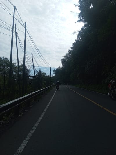 The image depicts a road in a forested area. The road is dark asphalt with a yellow line dividing the lanes and a white line marking the edge. On the left side of the road is a guardrail with vegetation growing behind it, and utility poles with wires extending across the sky. On the right side of the road is a dense forest. There are several people on motorbikes on the road. The sky is cloudy. The image is purportedly of Khe Luang, Laos, but that cannot be forensically verified solely from the content of the image.