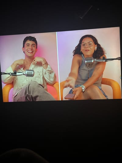 The image shows a split-screen view of two individuals likely participating in a podcast or similar recording session. The background appears to be a studio setting with a gradient pink and white backdrop. The top half shows a woman with curly hair, wearing glasses, a grey top, and denim shorts. She has a tattoo on her arm and is holding a phone. A vintage microphone is positioned in front of her. The bottom half shows a man with short dark hair, wearing a white patterned shirt and grey pants. He is smiling and also has a vintage microphone in front of him. They are both seated on yellow chairs. The image was captured indoors, likely on a screen, with dark surroundings obscuring the edges. There is no information about the location in the city of Parroquia Ocumare, Venezuela.