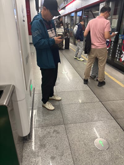 A subway platform scene shows several people waiting. One man in a pink shirt and khaki pants is looking at his phone. Another person, wearing a blue jacket and a black baseball cap, is also looking at a phone. A green arrow points the direction of travel on the platform floor. The subway car door is partially visible.