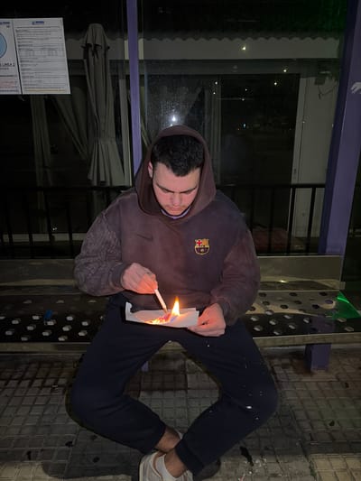 A male, approximately 18-25 years old, with dark hair, is seated on a perforated metal bench within a purple-framed public shelter at night in Grau i Platja, Spain. He wears a dark brown hooded sweatshirt featuring a Nike swoosh and an FC Barcelona crest, dark blue sweatpants with white splatters, and light-colored sneakers. The subject is intently focused downwards, holding a white rectangular piece of paper or cardstock in his left hand, which is actively burning with a significant flame and glowing embers. In his right hand, he holds a thin, white, cylindrical object with an orange-glowing tip, positioning it over the burning paper, indicating an ignition or fuel application action. A blue disposable lighter rests on the bench to his left. Through the shelter's glass panels, a dark outdoor environment with distant lights and a folded beige umbrella is visible. A white sign, partially obscured, displaying "LA MARINA" and "LÍNEA 2 GRAU I PLATJA" is affixed to the shelter structure in the upper left background. The ground is tiled.