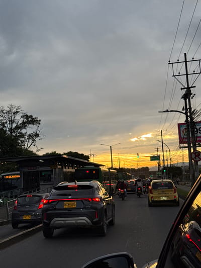The image depicts an urban traffic scene during sunset in the Area Metropolitana Centro Occidente (AMCO), Colombia, specifically near the "1 30 DE AGOSTO" bus stop. The perspective is from inside a stationary vehicle, showing a portion of its dashboard reflection and the passenger side mirror. A multi-lane asphalt road is congested with vehicles.
In the foreground, a dark grey SUV with license plate NXM-505 displays illuminated brake lights, facing away from the camera. Immediately to its left, a grey hatchback with license plate TMP-756 also shows illuminated brake lights. Further down the road in the right lane, a yellow taxi marked "SERVICIO PÚBLICO" is visible. Multiple motorcycles with riders, some wearing helmets, are interspersed among the cars.
On the left side of the road, a bus stop structure with a shelter and ramp is present. Two green public buses are visible, one at the bus stop displaying "1 30 DE AGOSTO" and route "2". Utility poles laden with numerous wires and streetlights line the right side of the road. A large commercial billboard is partially visible on the right. Traffic lights are present, with at least one displaying a green signal. The sky is partly cloudy, with a prominent orange and yellow sunset on the horizon to the right. Distant geographical features, possibly hills or mountains, are faintly discernible in the background.