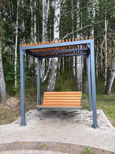 The image depicts an outdoor scene featuring a modern metal and wood pergola with a built-in bench. The pergola has a dark grey metal frame and a slatted wooden seat. It is positioned on a paved pathway made of grey interlocking bricks, which is bordered by grass and dirt. Behind the pergola, a dense forest of birch trees with characteristic white bark and green foliage is visible. The trees are in the foreground, creating a natural backdrop for the man-made structure.

The scene appears to be set during the daytime, with natural light illuminating the area. The weather seems overcast or possibly after rain, as the paving stones appear wet in some areas and there are some fallen leaves on the ground, suggesting an autumn season. The location is identified as Zarechny, Russia, which aligns with the presence of birch trees often found in this region.

There are no people visible in the image, and no specific activity or event is taking place. The focus is on the structure and its natural surroundings. The overall impression is one of a quiet, perhaps recently constructed, public seating area within a park or forest setting. The construction of the pergola suggests it is a place for relaxation or shelter. No visible text is present in the image.