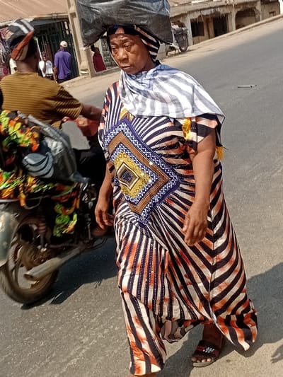An older woman in a patterned dress sits sideways on a motorcycle. Her dress has striped sections of white and blue, plus orange and brown patterns. A building is visible in the background. Additional people and objects fill the space around the motorcycle. The background suggests a street or market setting. The photo seems to have been taken at an angle, adding a unique perspective.