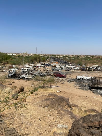 The image shows a large number of burned-out vehicles in Tayba Alhasanab, Sudan, located at coordinates 15.36 latitude and 32.528 longitude. The vehicles are scattered across a dirt field, and some are partially burned. The scene appears to be aftermath of a fire, suggesting the vehicles were intentionally destroyed. The surrounding environment is dry and dusty, with a few trees in the distance.