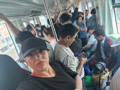 The image is a selfie taken inside a crowded train, likely a metro or subway car. The photographer is a light-skinned woman wearing a black baseball cap and a black shirt.
Multiple people are visible in the background, many holding onto the handrails. The passengers appear to be of various ethnicities, and ages. The train car is brightly lit with white walls.
Based on the urban scene visible through the windows, the image may have been taken in La Victoria, Peru.