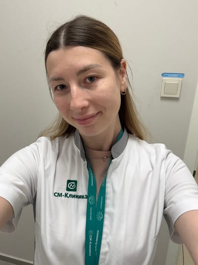 A young woman, appearing to be in her early twenties, takes a selfie in a white medical uniform. She has light skin with freckles, blue eyes, and medium-length brown hair. She is wearing a necklace with a small charm. A green lanyard with the logo "СМ-Клиника" is visible around her neck, suggesting she works at this clinic. The background appears to be an indoor hallway, with a light gray wall and a light switch plate visible on the right. There is a blue sign above the light switch with Russian text. No specific location in Borodino, Russia, can be determined from the image.