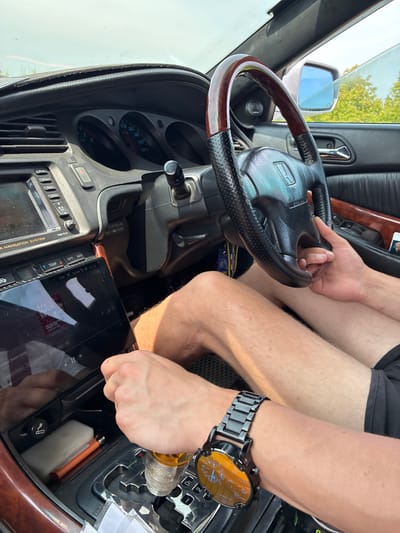 The image shows the interior of a car. A person's hands are visible. One hand is on the steering wheel, which has a wooden trim. The other hand is near the gear shift. A tablet is mounted on the dashboard, displaying what appears to be a music interface. The car's dashboard has a navigation system and various buttons. The scene suggests the car is either parked or moving slowly, as indicated by the ambient lighting and the casual posture of the occupant. The location is identified as Chernigovka, Russia, though no specific landmarks within the city are visible in the image.