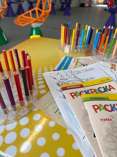 Here is a forensic description of the image:
The image presents an eye-level shot of a table in what appears to be a creative or activity-oriented space. The table is covered with a bright yellow sheet featuring white polka dots. Several coloring books titled "PACKPACHA" in Russian, stacked on top of each other, are visible. Each book displays different subtitles; "MYA," "MУФЛОН," and "ПУМА" are readable.
Two containers filled with colored pencils are placed on the table. The pencils range in colors, including shades of red, brown, yellow, purple, blue, and green.
Behind the containers, there is a construction toy.
The location in the city of Синьково, Russia is implied by the Cyrillic text on the books.