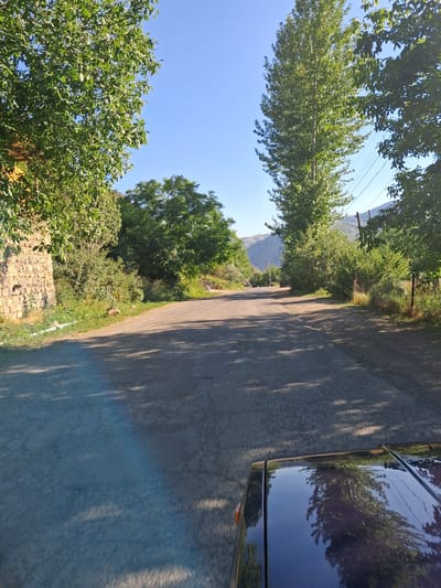 This media file shows a view from a vehicle driving on a paved road through a rural area. The scene is dominated by lush green trees and vegetation, with a clear blue sky above. The road is shaded by trees, casting dappled shadows on the asphalt. On the right side of the image, the side and roof of a dark-colored vehicle are visible, reflecting the surrounding trees and sky.
In the background, mountains can be seen in the distance under the bright sky. The overall impression is of a sunny day in a picturesque, natural setting. The time of day appears to be morning or afternoon, given the angle of the sun and the presence of strong shadows. There are no people or explicit text visible in the image. The setting suggests a drive through a countryside location, possibly in a mountainous region, consistent with the provided location context of Shatin, Armenia. The image captures a serene and tranquil moment of travel through a green landscape.