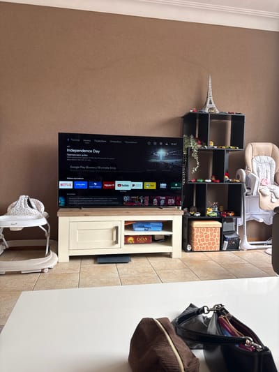The image shows a living room scene, likely in Veghel, Netherlands. A large television displays a streaming service interface, showcasing movie titles like "Independence Day" and various apps such as Netflix, Prime Video, and YouTube. The TV is positioned on a black shelving unit filled with small toy cars and decorative items. To the left of the shelves, a miniature Eiffel Tower figurine stands tall.
Beside the TV is a light-colored wooden cabinet with a drawer. On top of this cabinet, several board games are visible, including "CATAN." A black handbag rests on a white surface to the right of the cabinet, with a brown fabric pouch beside it.
In the background, a baby bouncer is set up, suggesting the presence of a child. The floor is a warm brown carpet, and a tiled wall is visible behind the cabinet. The lighting suggests it is daytime, though no specific time is explicitly indicated beyond the clock on the TV screen showing 10:47. The overall atmosphere appears to be domestic and relaxed. The visible text is primarily from the television screen, displaying app names, movie titles, and user interface elements in Bulgarian.