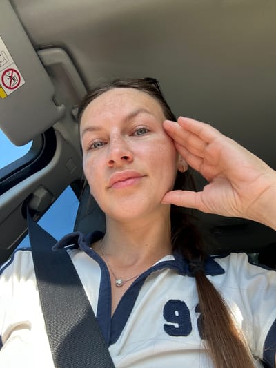 A woman is taking a selfie in a car. She is wearing a white polo shirt with blue trim and a number 9 on the front. She has on a silver necklace with a pendant. Her hand is resting on her face. The sun visor in the car is down, and there is a seatbelt visible. The background shows a clear blue sky and part of the car's interior. The image was taken in Vrela, Montenegro.