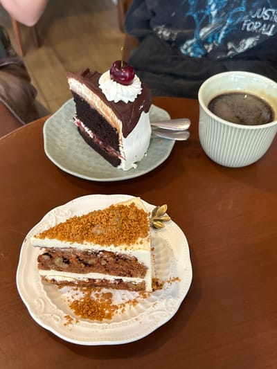 The image displays a close-up, slightly overhead view of two slices of cake and a cup of coffee on a dark brown wooden table, situated in an indoor setting in Chiang Dao, Thailand.

In the upper-middle portion of the frame, a slice of chocolate cake rests on an irregular, light green ceramic plate. The cake consists of several layers: a dark brown chocolate sponge, a lighter brown mousse or cream layer, and a white cream layer, with a thin reddish fruit or jam layer visible near the bottom. The top is covered in a smooth, dark chocolate ganache, topped with a dollop of white whipped cream, and garnished with a single red cherry featuring a green stem. Two silver-colored dessert spoons are positioned on the plate next to the cake.

To the right of the chocolate cake, a light green ceramic cup with vertical ridges contains a dark, presumably black, coffee.

In the lower-middle portion of the frame, a slice of carrot cake is presented on a white, scalloped-edge ceramic plate with an embossed pattern. This cake has distinct layers of a reddish-brown sponge, visibly containing walnuts and possibly other inclusions, alternating with layers of white cream cheese frosting. The top is heavily covered with a coarse, light brown crumb topping, potentially biscoff or graham cracker crumbs, and a small, translucent, light-colored garnish. A small, decorative gold-colored item shaped like a leaf is partially visible on the plate's edge.

Portions of two individuals are visible in the blurred background. In the upper left quadrant, a light-skinned arm and shoulder are present. In the upper right quadrant, a person wears a dark t-shirt with a distressed blue and white graphic design that includes the text "JUSTICE FOR ALL". The immediate table surface is a consistent dark brown.