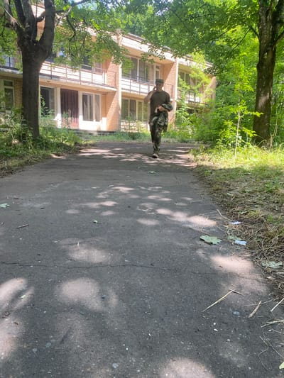 A man in camouflage pants and a dark green t-shirt, holding a jacket in his left arm and speaking on a mobile phone in his right, walks along an asphalt path. The path is lined with dry grass and leaves on the right side. A large tree with green foliage is on the left, and a three-story brick building with balconies and numerous windows is visible in the background. The scene appears to be outdoors, possibly in a park or abandoned area, on a sunny day with dappled sunlight. The environment suggests a suburban or rural setting, consistent with the outskirts of Shelkovo, Russia.