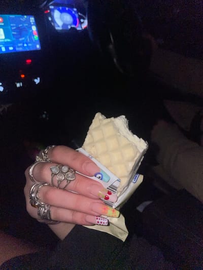The image captures the interior of a vehicle at night. In the foreground, a left hand holds a partially consumed white ice cream bar. The bar has a waffle-patterned surface, a creamy white interior, and a bite taken from its upper right corner, remaining partly in its white and blue packaging. The hand is adorned with multiple silver-toned rings on the index, middle, ring, and pinky fingers. The index finger features a large ring with five clear or translucent stones. The fingernails are long and manicured, displaying distinct designs: an "evil eye" on the index finger, red cherries on the middle finger, yellow flowers on the ring finger, and red polka dots on a white base on the pinky finger. In the background, the vehicle's dashboard is partially visible with illuminated screens. A left screen displays a map or navigation interface, while a right screen shows the number "78," indicative of a speedometer. A light-colored limb is visible in the upper right background. The specific location within Ndlambe Local Municipality, South Africa, cannot be determined from the provided visual information.
