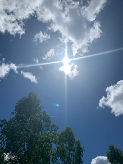 This is a ground-level, low-angle shot of a bright blue sky with scattered white clouds and a prominent sunburst. The sun is positioned near the top center of the frame, casting lens flares that create streaks of light and a subtle rainbow effect. The upper portion of a deciduous tree with green foliage occupies the bottom third of the image, extending upwards from the left side. A small, out-of-focus blue light artifact is visible in the sky to the right of the sunburst. There are no people, animals, or man-made structures clearly identifiable in the scene, suggesting an outdoor, natural environment. Given the provided context of Vodoratsk, Russia, this image likely depicts a clear, sunny day outdoors in a park or natural area within or near the city.