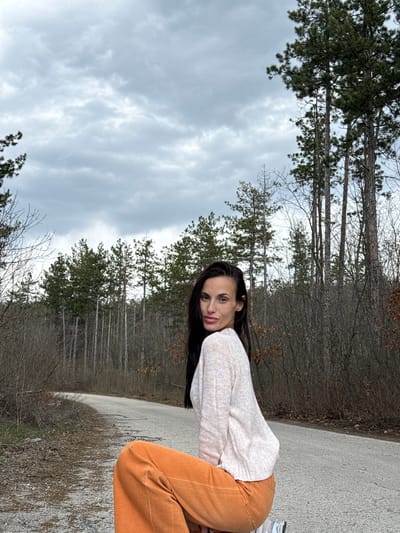 The image displays an outdoor scene located in Radishevo, Bulgaria. A single female subject is positioned on a paved road within a wooded environment under an overcast, gray sky. The woman has long, dark hair and is wearing a light-colored, long-sleeved knitted sweater, bright orange trousers, and white sneakers with gray accents. She is in a crouching or kneeling stance on the left side of the road, with her body angled slightly right, and her face turned towards the camera, maintaining direct eye contact. The road, an asphalt surface appearing somewhat worn, curves to the left in the background. It is flanked by dense vegetation, primarily consisting of tall coniferous trees with green needles and numerous deciduous trees with bare branches, indicating a cooler season.
