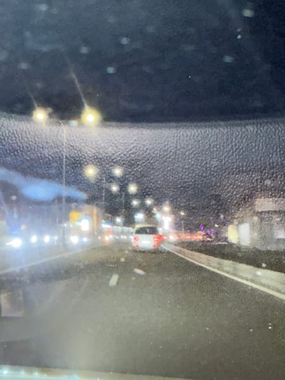 This image depicts a night-time view from inside a vehicle traveling on a road, likely a highway, in Parakar, Armenia. The foreground shows the interior of the car's windshield, which is speckled with water droplets or dirt, creating a blurry effect. The road ahead is visible, with lane markings illuminated by streetlights and vehicle headlights. Several cars are on the road, some with their headlights or taillights on, suggesting active traffic. In the background, there are buildings and streetlights, indicating an urban or suburban environment. The overall lighting is dim, characteristic of nighttime, with sources of light creating a hazy or diffused illumination. The presence of streetlights and buildings suggests infrastructure typical of a city or town.