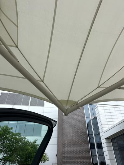A low-angle shot captures the underside of a light-colored fabric awning supported by a metal frame. The awning is segmented into panels by darker seams and extends over a modern building. To the left, the open door of a vehicle reveals a green tree and a building with horizontal vents and large windows. The building features a facade of white panels and dark brick.