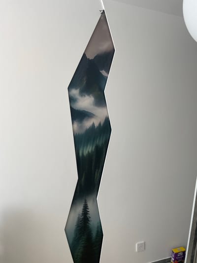 The image displays a vertical section of a wall featuring a scenic mural of a misty forest with mountains. The mural is revealed through a jagged, vertical cutout in the otherwise plain white wall. In the lower right corner of the image, a white electrical outlet is visible on the wall, and a yellow and purple box, likely containing wallpaper paste, is situated on the floor near a gray baseboard. A portion of a white ceiling light fixture is visible in the upper right corner. The scene appears to be indoors, possibly in a room under renovation or decoration, as suggested by the wallpaper paste. The location of this scene within the city of Meaux, France, is not discernible from the provided image.