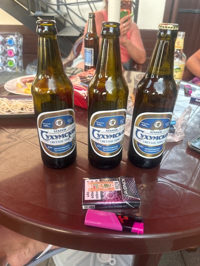 Three bottles of Sukhunsky Svetloe Pivo (light beer) are on a brown, circular table. The bottles are dark green glass with labels that are primarily blue and white, featuring text and images related to the beer. One bottle is missing its cap, while another has a gold crimped cap. To the right of the beer bottles, there is a pack of cigarettes with black and purple packaging and a pink lighter. In the background, a person in a pink top is partially visible, and another person's legs are visible at the bottom of the frame. A plate with food and a plastic container are also on the table. The scene appears to be outdoors, possibly in a cafe or restaurant setting, with an Abkhazian flag visible on the cigarette pack.