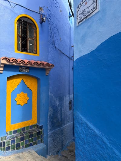 The image displays a narrow, stepped alleyway in Chefchaouen, Morocco, identified by a white street sign reading "Calle Sidi Buchuka" in Latin script and Arabic above it. On the left, a building painted in a vibrant blue features an arched window with a bright yellow frame and an ornate black wrought-iron grille, partially open. An external light fixture is mounted above the window. Below the window, a blue doorway with an elaborate yellow arched frame and a central yellow starburst motif is visible. A terracotta-colored ridged canopy with white supports is positioned above the door, with a small white plaque featuring Arabic calligraphy underneath it. The lower section of the doorway is tiled with a pattern of dark green and blue squares. The adjacent wall on the right is also painted blue, distinctly segmented into a lighter sky-blue upper portion and a darker, undulating royal blue lower section. Stone steps descend into the foreground of the alley. No individuals, animals, or active elements are present in the scene.