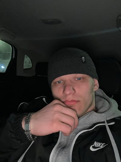 The image displays a male subject, appearing young adult, captured in a close-up, frontal portrait within a vehicle interior. The subject possesses blue eyes and maintains a neutral expression, with his left hand resting beneath his chin. He wears a dark grey ribbed knit beanie with a discernible rectangular brand tag on the front, a grey hooded sweatshirt, and a black athletic jacket featuring a white Nike logo on the right chest. A silver-toned metal link bracelet is present on the subject's left wrist. The vehicle's cabin is characterized by low light conditions and dark upholstery. Exterior elements are visible through the rear-left window, consisting of blurred artificial lights and faint architectural outlines, indicative of a nighttime setting. The precise geographical location within Dobryanka, Russia, is not ascertainable from the visual evidence.