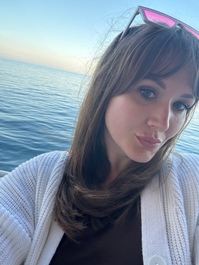 A woman with brown hair and bangs, wearing pink sunglasses, is posing for a selfie. She is puckering her lips. She is wearing a white sweater with a button. Behind her is the sea. The location is likely in or near Гаспринский поселковый совет, Ukraine, given the possible outdoor and sea background.