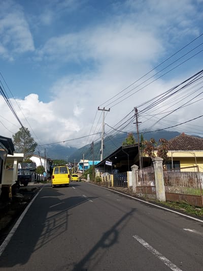 The image shows a street in a residential area. Two yellow vans are driving on the asphalt road. Buildings line both sides of the street, with power lines overhead. Mountains are visible in the background, partially obscured by clouds.