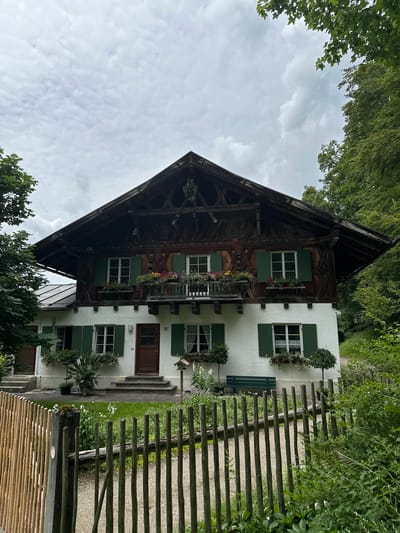 This is a vertical, eye-level, full shot of a charming, traditional Bavarian house. The house has a white facade with green shutters on its windows, and a steeply pitched, dark wooden roof. Decorative wooden carvings adorn the gable, and flower boxes filled with colorful blooms are placed beneath several windows and on the balcony. A wooden fence, made of rough-hewn logs, runs along the right side of the image, separating the house from a gravel path. Lush green trees and foliage surround the property. The sky is overcast with gray clouds, suggesting a cloudy or possibly rainy day. The scene is peaceful and idyllic, evoking a sense of rural tranquility. There is a wooden bench visible on the left side of the fence. A small plaque with the number "10" is visible near the front door. The overall impression is of a well-maintained, picturesque home in a natural setting.