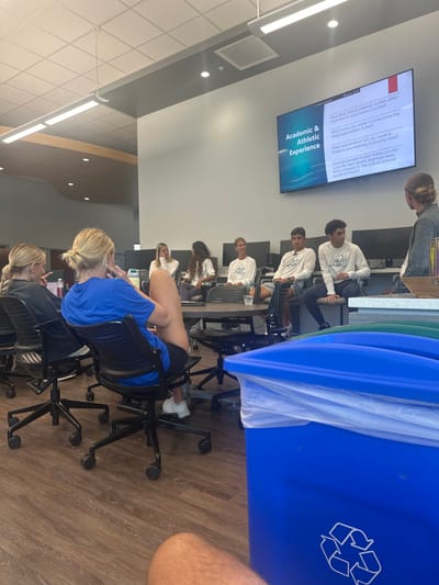 The image depicts an indoor panel discussion or seminar occurring in a modern room located in Miromar Lakes, United States. The room features light grey walls, a white drop ceiling with linear LED light fixtures, and light brown wood-grain laminate flooring.

On the back wall, a large flat-screen television displays a presentation slide titled "Academic & Athletic Experience." Visible text on the screen includes questions such as "How does the academic system differ from that in your home country?" and "What have you done to overcome the language barrier, if any?" It also mentions "adjustments did you have to make in your training or playing style?" and references "the culture around sports at FGCU."

A dark, round table is positioned centrally, accommodating a panel of six individuals seated in black wheeled office chairs. Five panelists—three females and two males—wear white long-sleeved shirts with "FGCU" branding. The sixth panelist, a female, wears a plain white long-sleeved shirt. Clear drinking glasses are on the table. To the far right, a male with light brown hair styled in a bun, wearing a grey long-sleeved shirt, stands facing the panel, appearing to moderate or present. Behind the panel, multiple computer monitors are visible on desks lining the back wall.

To the left, two blonde females are seated in black wheeled office chairs, observing the scene. One, with her hair in a ponytail, faces left. The other, wearing a blue t-shirt, black shorts, and white sneakers, has her hair in a bun and leans back. In the immediate foreground on the right, a blue recycling bin with a clear plastic liner and a white recycling symbol is prominent, partially obscuring a green recycling bin behind it. The photograph is taken from a seated individual's perspective, with bare legs visible at the bottom center.