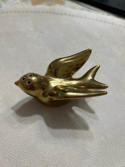 A close-up, eye-level, full shot shows a small, hollow, brass bird sculpture with its wings outstretched, appearing to be in flight. The bird is resting on a textured, light-colored surface, casting a distinct shadow to its right. The sculpture has four small holes on its head, suggesting it might be a salt or pepper shaker. The bird is facing left, with its beak pointed slightly upwards. The surface beneath the bird is a light beige with a subtle, pebbled texture. The background is out of focus, showing a faint pattern of light and dark colors. There are no people visible in the image. The scene is indoors.