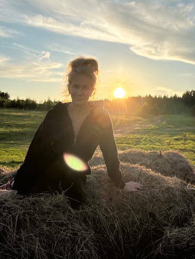 An outdoor portrait captures a woman during sunset in a rural area, likely near Стеклянный, Russia. The woman, with fair hair styled in an updo and a gentle smile, is positioned in the foreground, illuminated by the warm, golden light of the setting sun. She is wearing a dark, possibly zipped, top and her left hand is casually resting on a large, round hay bale to her right.

The sun, a brilliant, circular orb, is low in the sky directly behind her, creating a strong backlighting effect that silhouettes her figure and highlights the edges of her hair. A prominent lens flare, appearing as a bright, circular glow with rainbow-like rings, is visible over her right side.

The background features a verdant field or pasture stretching towards a distant line of trees or a forest along the horizon. The sky is bright with soft, wispy white clouds scattered across a light blue expanse, indicative of clear weather. The combination of the golden hour light, the natural setting, and the woman's relaxed expression evokes a sense of tranquility and contentment. There is no visible text in the image.