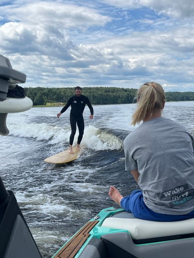 In Gromovo, Russia, a man in a black wetsuit is wake surfing on a lake. He stands on a light brown surfboard, riding the wave generated by a boat, which is partially visible in the foreground to the left. The man has a beard and a number "78" is visible on his wetsuit. In the background, a line of green trees borders the lake, under a sky filled with white, puffy clouds. A woman with blonde hair pulled back in a ponytail is seated in the boat, facing away from the camera, wearing a grey t-shirt that reads "Wake Surf Mafia". She is wearing blue sweatpants.