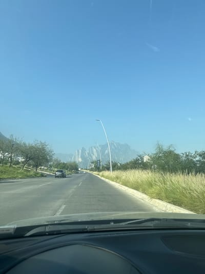 The image presents a forward view from the interior of a vehicle on an asphalt road in Ciudad Santa Catarina, Mexico, under a clear, light blue sky. The vehicle's dashboard and windshield wipers are visible in the lower portion of the frame. The road features at least two lanes in the direction of travel, marked by a white dashed line. A single dark-colored sedan is observed in the left lane ahead. The left side of the road includes green grass, scattered trees, and distant low-rise buildings. The right side is bordered by a concrete curb, beyond which lies a wide strip of tall, light green/yellowish feathery grass, followed by denser green trees and some structures. Two tall, white, curved streetlights stand on the right side of the roadway. In the far background, large, jagged, light grey mountains are prominent against the horizon. No individuals are visible.