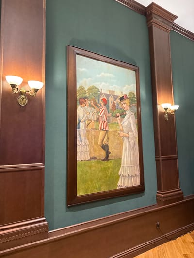 The image depicts a large, framed painting hung on a dark teal wall, flanked by dark wood columns. The painting itself is a scene of people playing tennis in what appears to be an outdoor setting with a house and trees in the background. The style of dress suggests a historical period, likely late 19th or early 20th century.

In the foreground of the painting, a woman in a white, ruffled dress and a straw hat stands holding a tennis racket and a glass of champagne. To her left, closer to the net, are three other figures, one in a striped tennis outfit, and two in white dresses, also holding rackets and glasses.

The wall on which the painting hangs is a deep, matte teal. On the left side of the painting, a dark wood column features a two-branched sconce with frosted glass shades, emitting a warm light. Similarly, on the right side, another dark wood column has an identical sconce. The base of the wall has a dark wood wainscoting with decorative molding.

The image was taken in a room with warm lighting, likely indoors, given the presence of the sconces. The overall impression is one of a formal interior space, perhaps a hall or a lounge, where art is displayed. The location in Zhukovka, Russia, is noted, but there are no visual cues within the image itself that confirm this specific location.