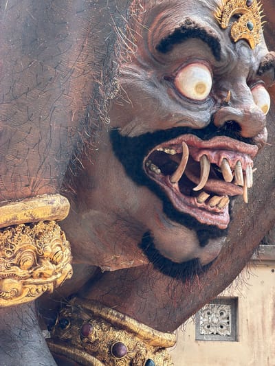 A large, detailed sculpture of a dark-skinned, demonic figure, likely an Ogoh-ogoh or similar cultural effigy, is depicted outdoors in Tegal Suci, Indonesia. The figure, seen from the mid-torso up and angled right, features heavily textured, dark brown skin with numerous individual dark and reddish-brown hair-like strands embedded in the surface.
The face is characterized by prominent, bulbous white eyes with small, dark central pupils and reddish irritation around the edges. A wide, menacing grin reveals a red mouth interior with numerous sharp, off-white fangs and tusks of varying lengths protruding from both upper and lower jaws, some long and curved. The nose is broad with two small tusks emerging near the nostrils. Dark, bushy eyebrows frame the eyes, and a prominent forehead is visible.
Elaborate gold ornamentation adorns the figure, including a multi-tiered crown-like structure on the forehead featuring intricate scrollwork and embedded green and purple cabochon-style stones. A golden collar or armlet with similar intricate patterns and red/purple stones is visible on the lower left portion of the sculpture. The overall impression is one of a formidable, powerful entity.
In the background, to the lower right, a segment of a light-colored (beige/off-white) building wall is visible. Set into this wall is a square, decorative white or light grey vent or panel featuring an ornate, symmetrical floral or geometric carving.