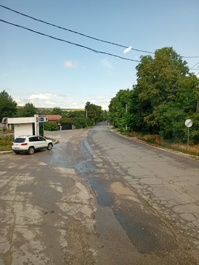 The image depicts a street scene in Trinca, Moldova. A white SUV is parked to the left of a bus stop shelter, which has the word "TRINCA" visible on it. A person in a pink shirt is walking away from the bus stop towards a fence. The road in the foreground is paved and appears to have some water or oil stains and cracks. Dense green trees line the right side of the road, and further in the distance, buildings and fields are visible. The sky is blue with some clouds. The scene appears to be a public space within the village.