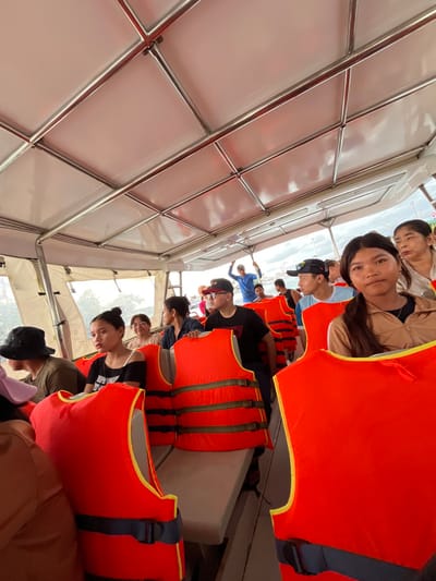 The image depicts the interior of a passenger boat in Sihanoukville, Cambodia, populated by approximately 15-20 individuals, primarily of Southeast Asian descent. The boat features a white overhead canopy supported by polished stainless steel framework, with light-colored bench seating arranged in rows. Numerous bright orange life vests, each with black horizontal straps and buckles, are folded and placed on the seats, serving as available safety equipment.
In the immediate foreground on the right, a young woman with long dark hair, wearing a brown jacket, is seated and looking directly towards the camera. Behind her and to the right, an older woman is partially visible. In the mid-ground left, a young woman with dark hair tied back, wearing a black off-the-shoulder top, is seated and looking left. A man in a black bucket hat and an olive-green shirt is seated in front of her. Other passengers are distributed throughout the cabin, including a man wearing a black cap and red-rimmed glasses, and another man in a black cap. Towards the stern of the boat, a man in a blue shirt and a light-colored cap stands, with his arms slightly raised, looking out over the water.
Through the open sides and stern of the vessel, calm water is visible, indicating the boat is either docked or in motion on a calm body of water. Distant land or port infrastructure, including what appears to be a pier or shoreline with utility poles, is faintly visible against a light sky, consistent with a coastal environment near Sihanoukville.