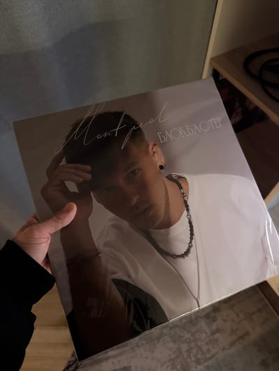 A photograph taken indoors shows a person holding a vinyl record album. The album cover features a man with short, dark hair wearing a white shirt and a black beaded necklace. The title "MONTREAL" is written vertically along the left side of the cover, followed by the word "БЛОКБАСТЕР" written vertically in Cyrillic. The album is partially covered in plastic wrap. The person holding the album is wearing a black long-sleeved shirt. The background includes a wooden shelf with visible objects and a wooden surface. The location is not specified in the image or its context.