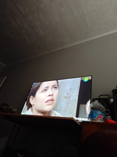 The image shows a woman on a television screen, appearing distressed or emotional, with a year "1943" displayed in the corner. A colorful logo with the Cyrillic text "МИР" is also visible in the upper right corner of the screen. The television is placed on a wooden desk, in front of a speaker labeled "SONY". There is a clear plastic bag containing some items on the desk next to the speaker, and a red object partially visible behind it. The background includes a textured ceiling and a wall. The scene appears to be indoors, likely a living room or entertainment area. The location is identified as Кашка-Суу, Kyrgyzstan.