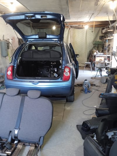 In an auto repair shop in Sukleya, Moldova, a blue Nissan Micra is parked with its hatchback and driver's side door open. The car's interior is partially visible, with the rear seats detached and placed in the foreground. Various tools and car parts are scattered around the shop, indicating ongoing repair work. A metal stool and a shelf unit with equipment are also present. The lighting suggests it is daytime.
