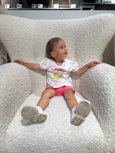 An overhead shot taken in Ustroń, Poland, shows a young child lying on their back on a large, textured, off-white fluffy chair or beanbag. The child has light brown hair and is wearing a white short-sleeved top with a partial graphic of a lemon and the word "LEMONADE," pink shorts, and white shoes.
The child's head is positioned towards the left side of the frame, and they are looking upwards and slightly to their left with a curious, slightly open-mouthed expression. Both arms are extended out to the sides, resting on the soft, shaggy material of the chair. Their legs are slightly bent, with the soles of their white shoes visible. Faint text, including "STEP-H" and other indistinguishable markings, can be seen on the sole of the right shoe.
In the background, to the left of the child, shelving units displaying various items are visible, suggesting an indoor commercial setting such as a furniture store or showroom. The overall scene conveys a sense of innocent curiosity as the child rests on the plush furniture. The lighting indicates it is daytime.
